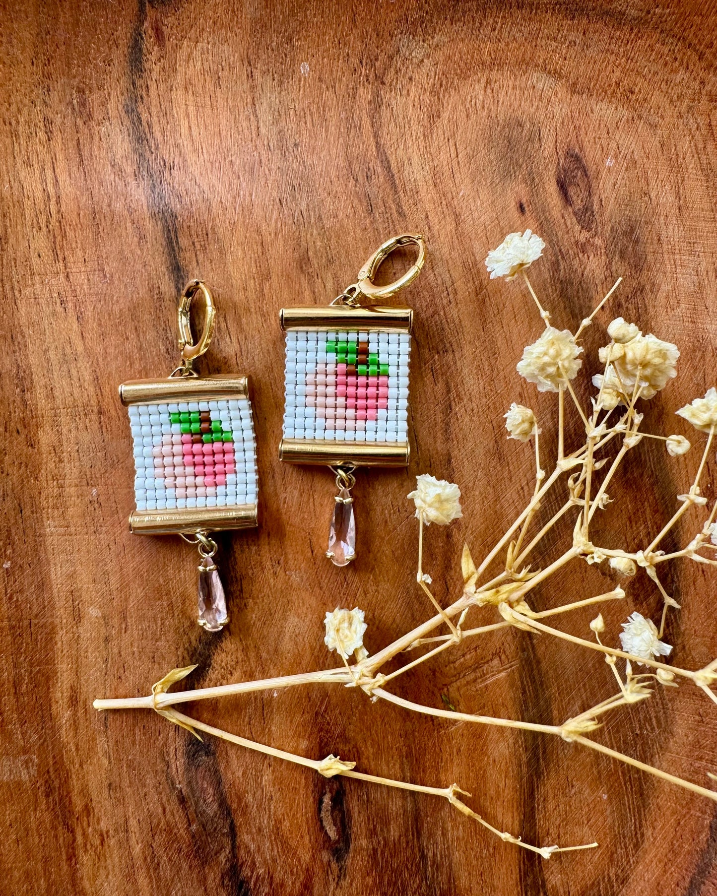 Two beaded earrings depicting peaches on a wooden surface with dried flowers.