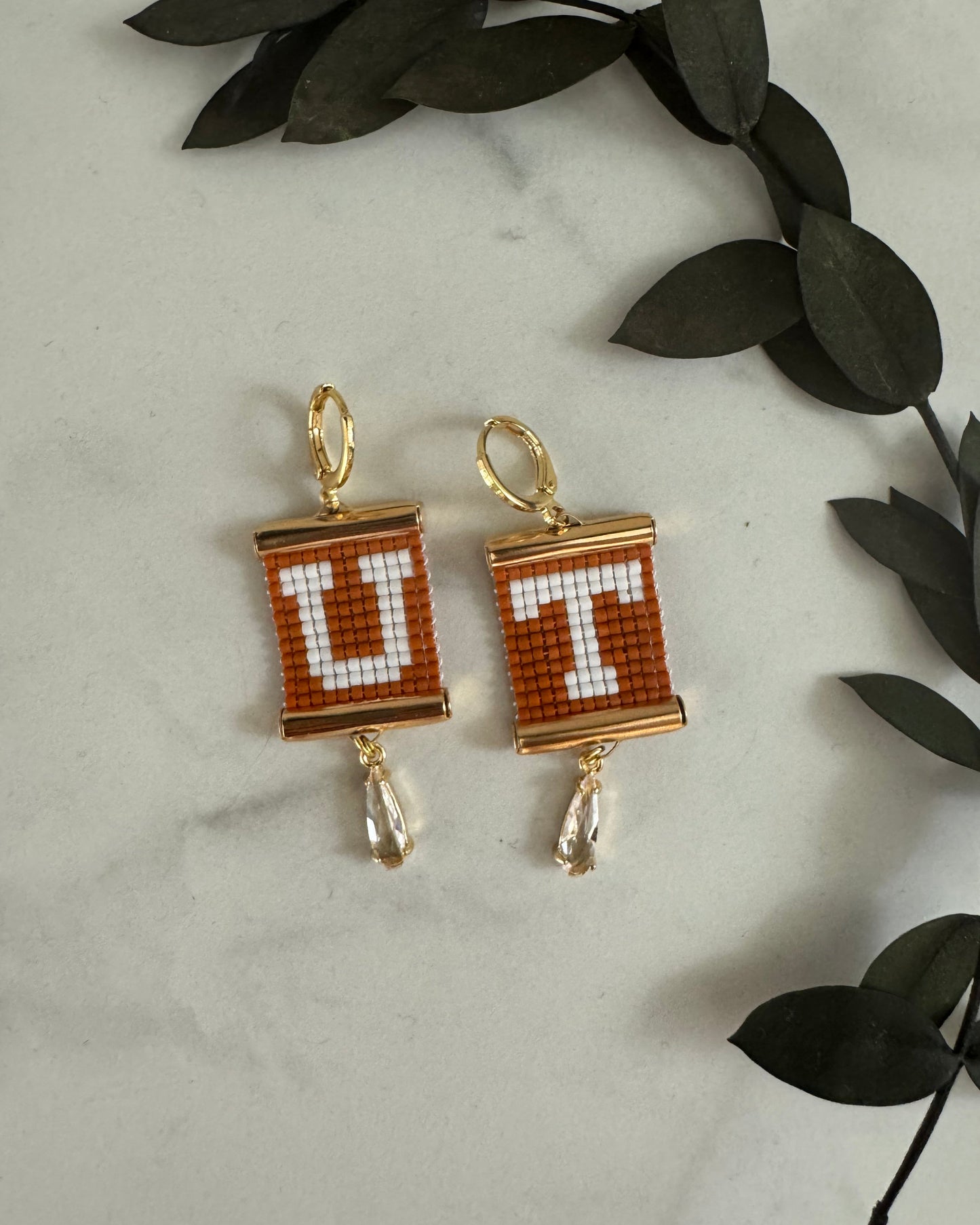 Two gold, white, and burnt orange beaded earrings with letters 'U' and 'T' on a marble surface with leaves.