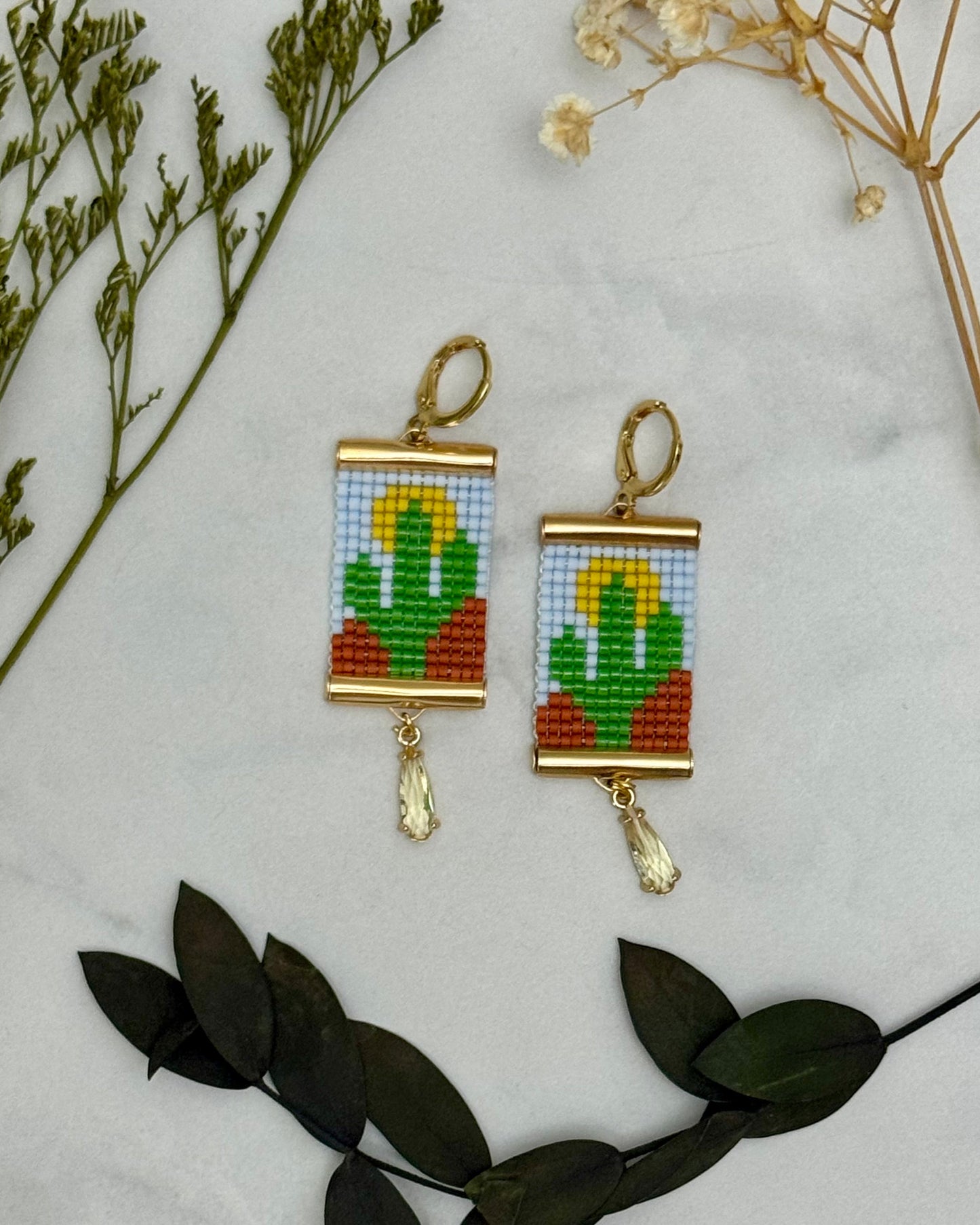 Beaded cactus earrings on a marble surface with leaves and flowers.