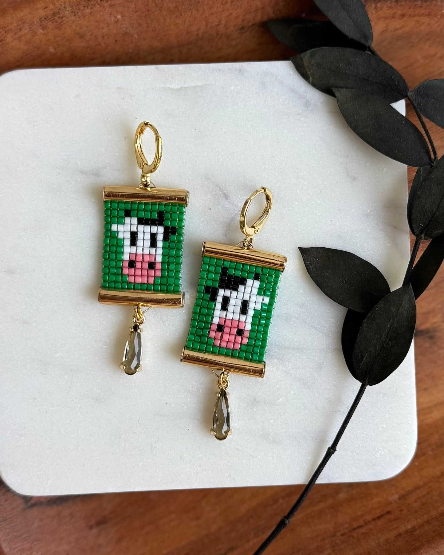 Pixelated cow earrings on a marble coaster with a wooden tray and leaf decoration.