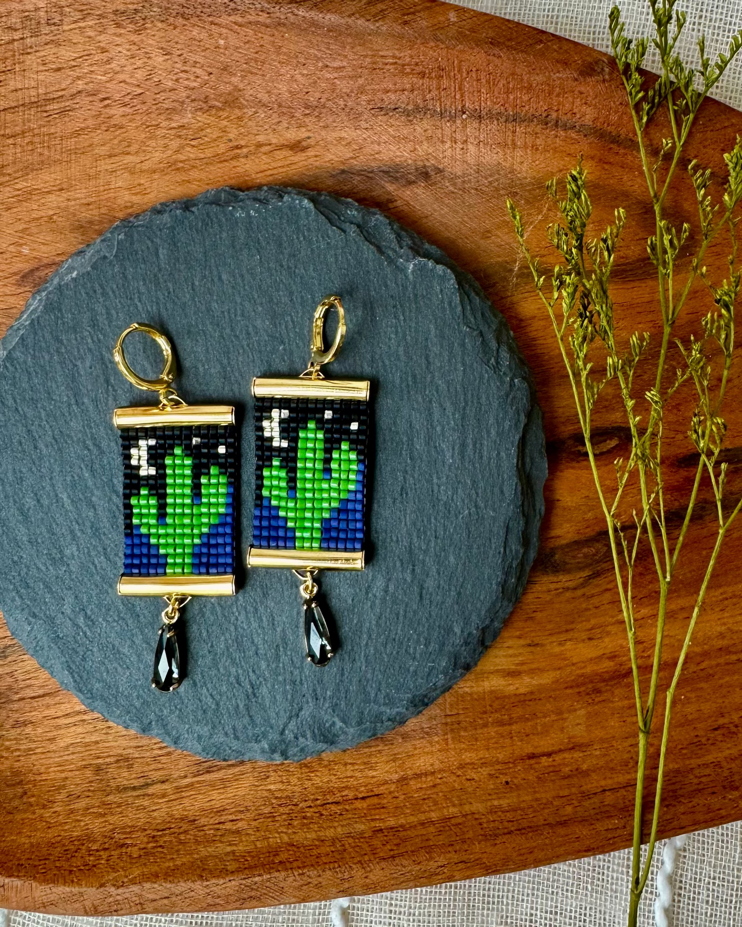 Pair of beaded earrings depicting saguaro cacti with the moon and stars on a slate stone with a wooden board and dried plants.