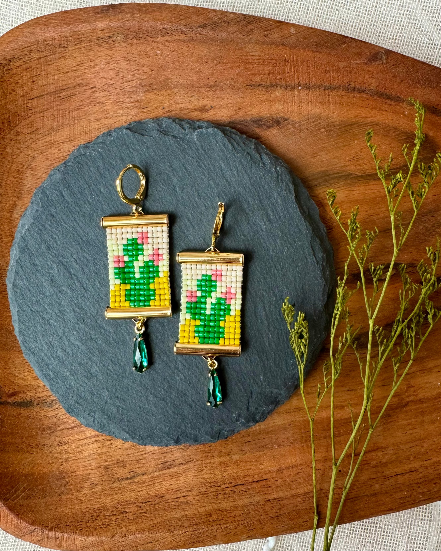Pair of earrings with cactus design on a slate stone, placed on a wooden surface with a plant.