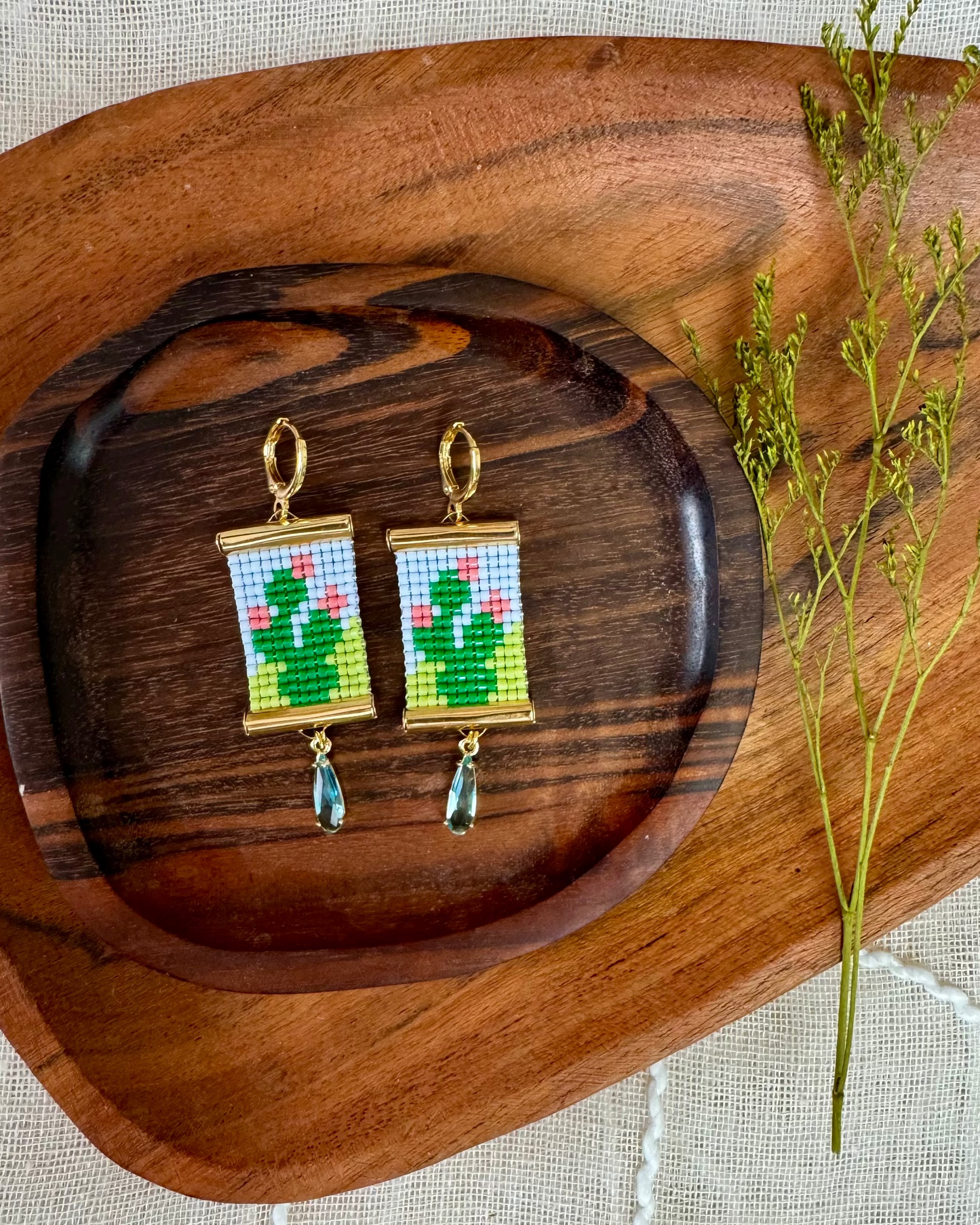 Pair of earrings with beaded prickly pear cactus pattern on a wooden tray with a plant stem.