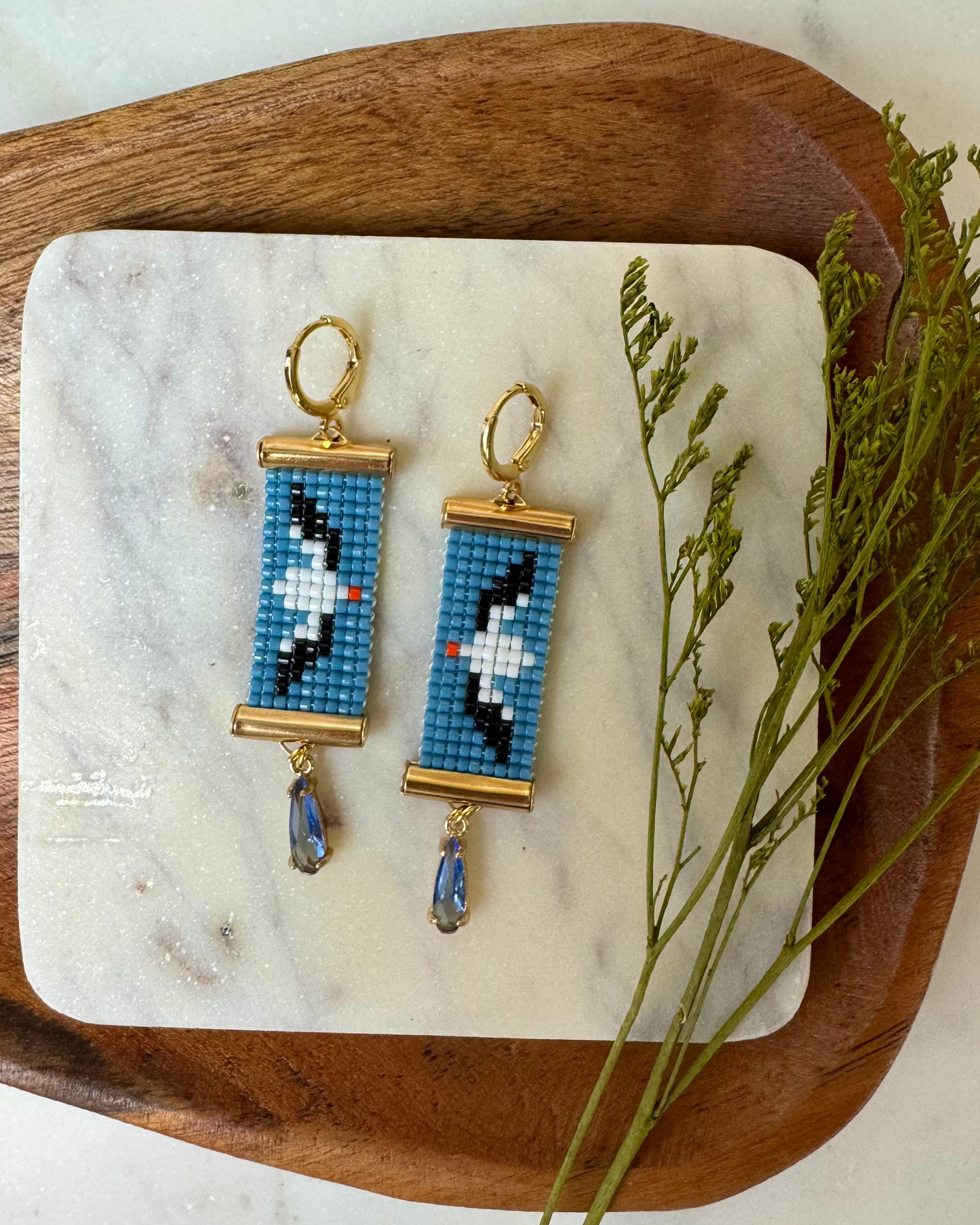 Pair of beaded earrings depicting albatross birds on a marble and wood tray with greenery.