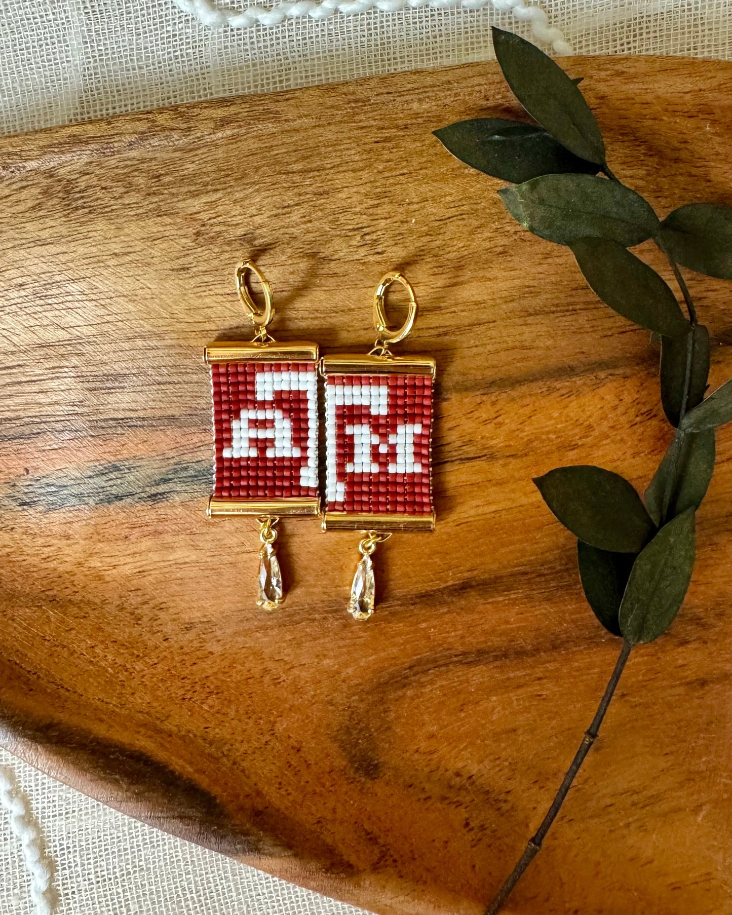 Beaded earrings with a 'A&M' design on a wooden surface with green leaves.