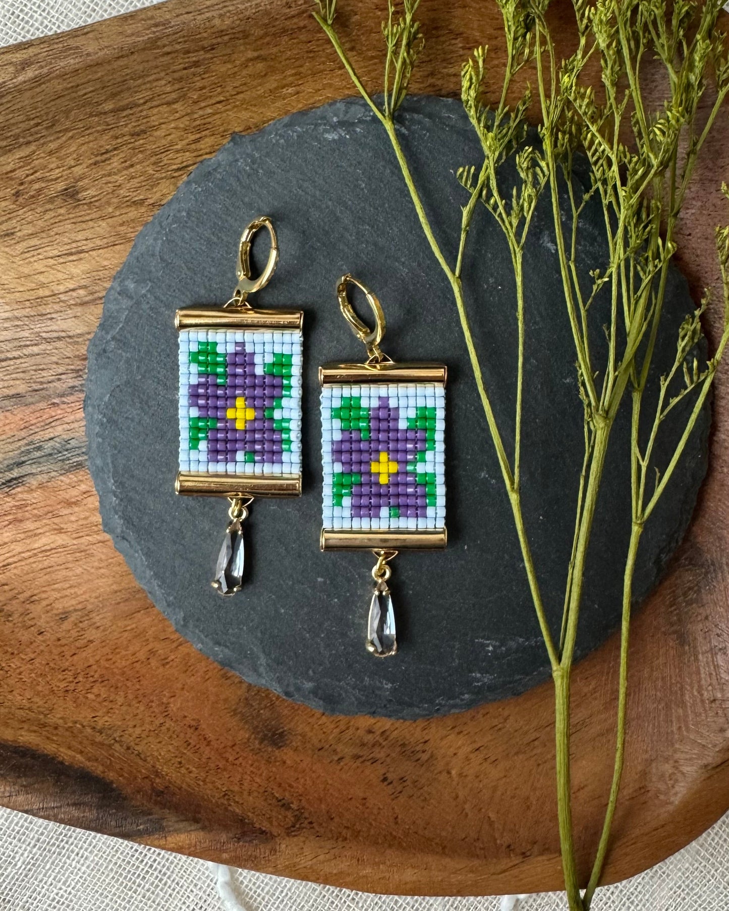 Pair of beaded earrings on a slate stone with a wooden base and dried plant.