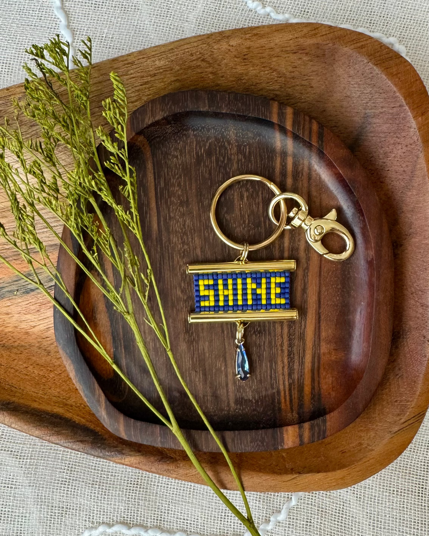 Beaded bag charm depicting the word 'SHINE' on a wooden tray with decorative elements