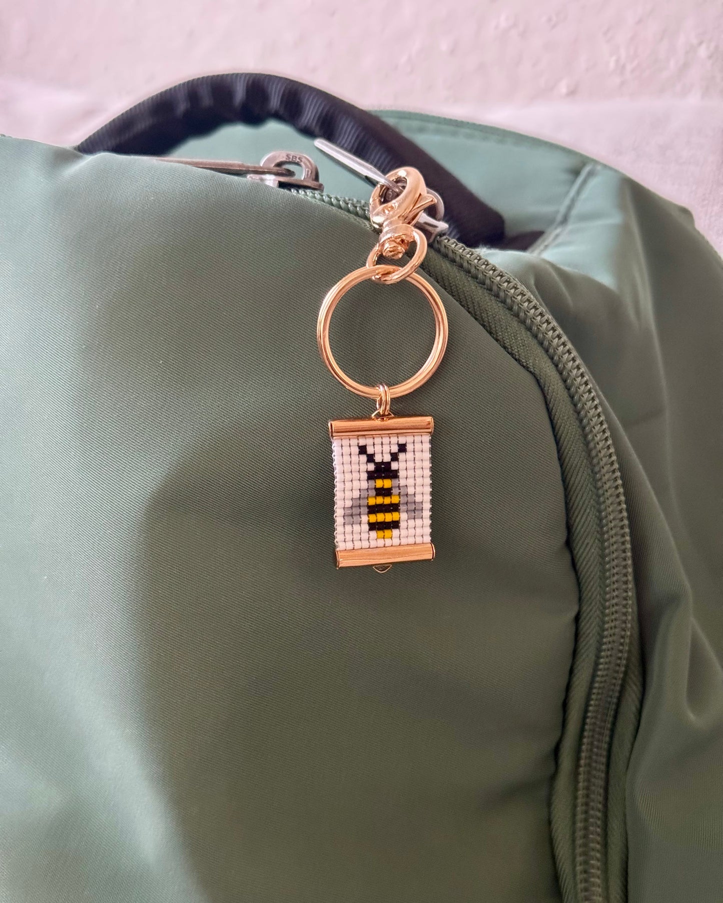 Woven beaded bag charm with a bee design attached to a green bag.