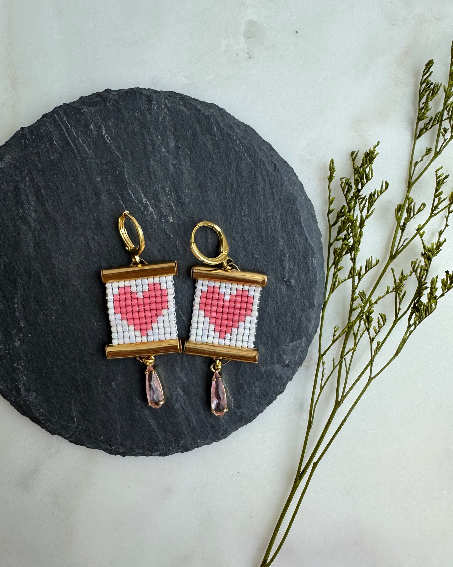 Pair of earrings with heart design on a round stone surface with dried plants.