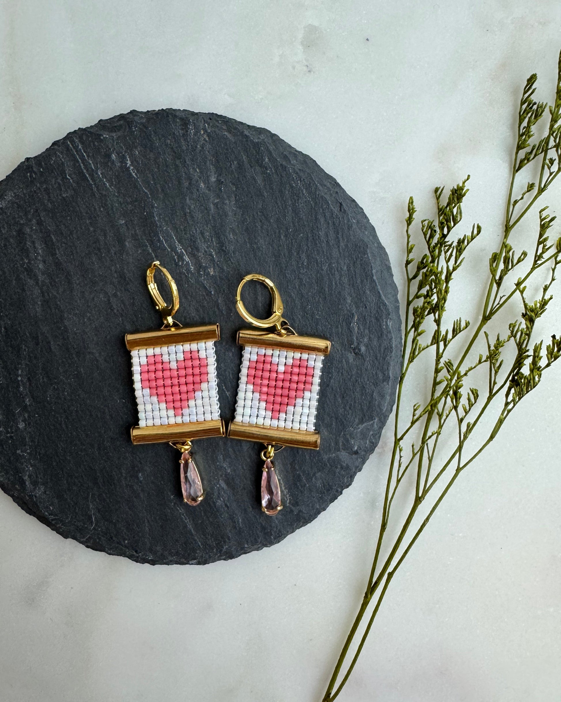 Pair of earrings with heart design on a round stone surface with dried plants.