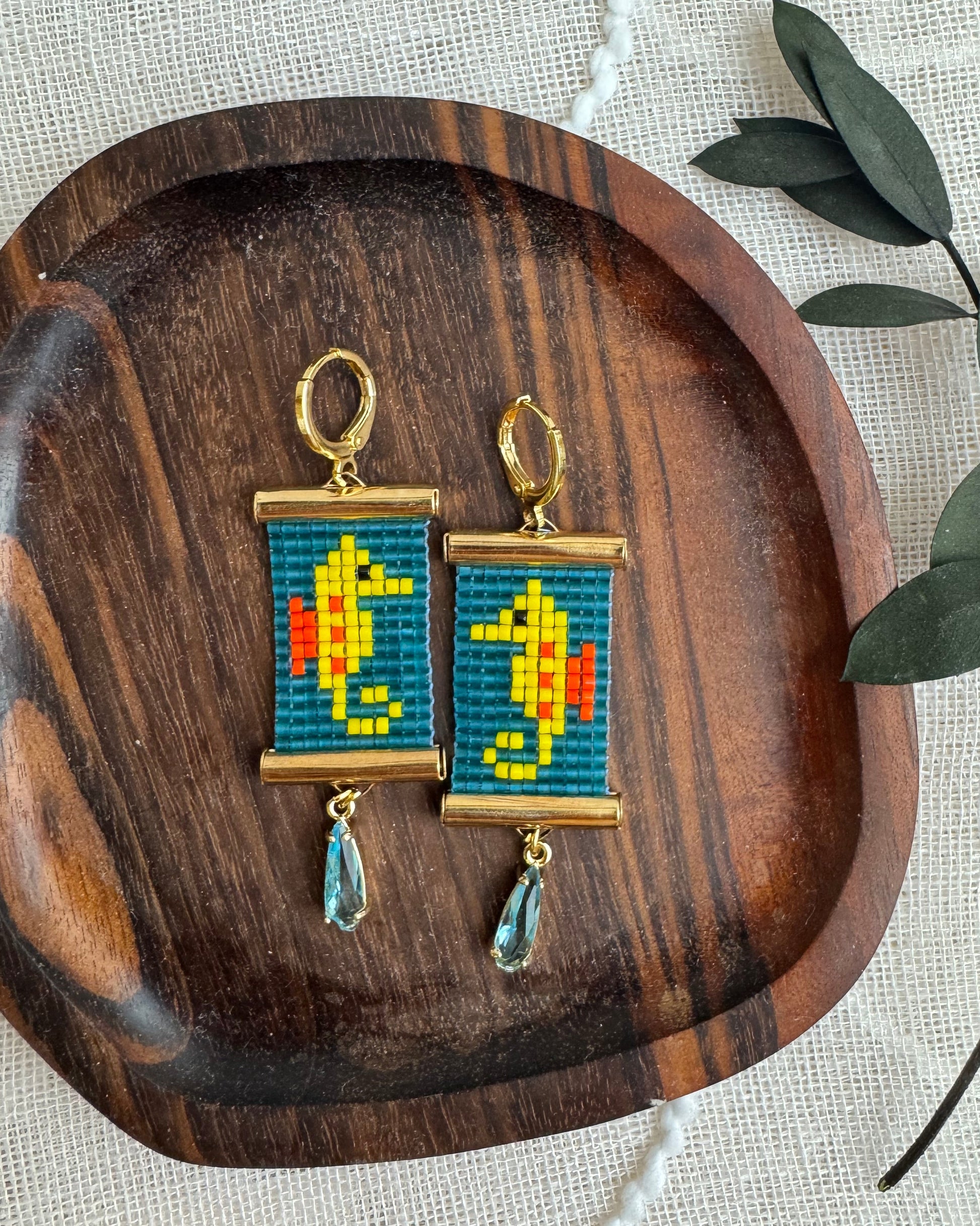 Two colorful earrings with seahorse designs on a wooden tray with a natural background