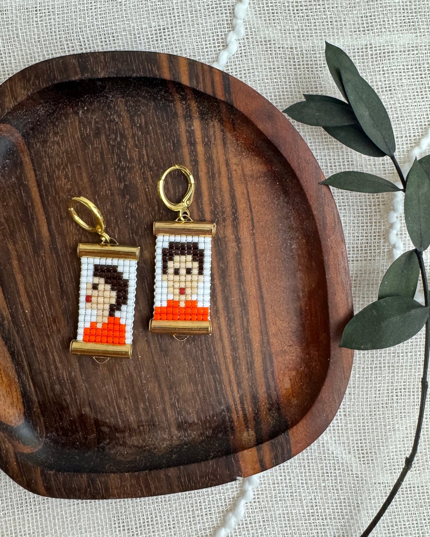 Pair of earrings with a pixelated design on a wooden tray with green leaves.