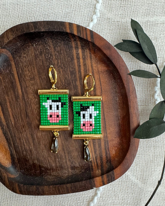 Pair of earrings with green background and cow design on a wooden tray.