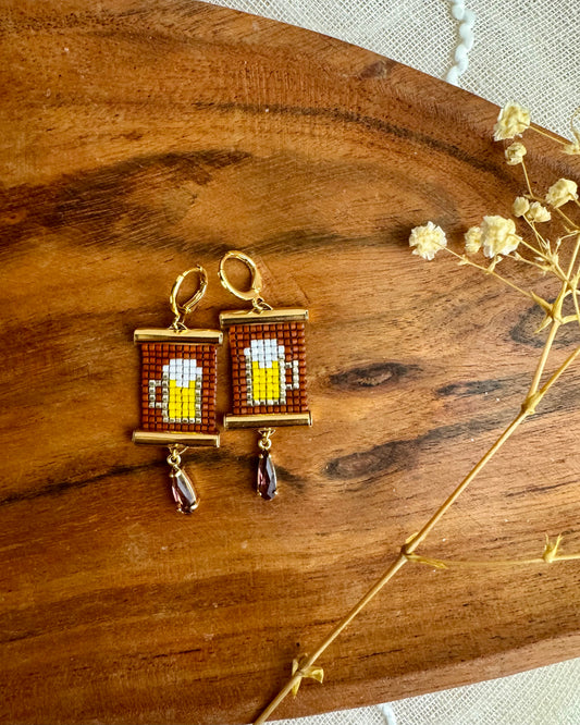 Pair of earrings with beer mug design on a wooden surface with dried flowers.