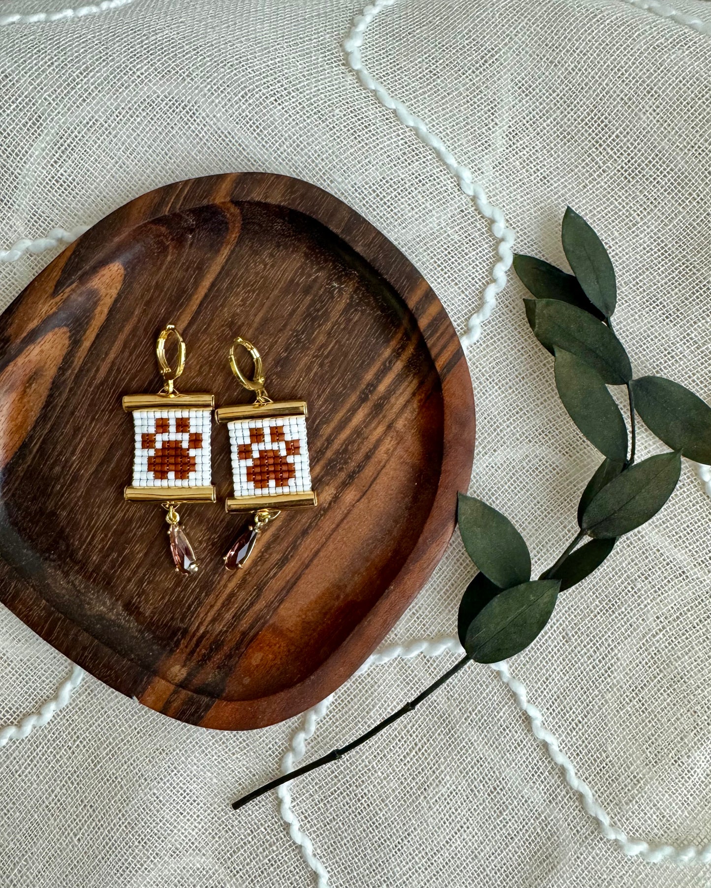 Pair of earrings on a wooden tray with a leafy branch on a textured fabric background