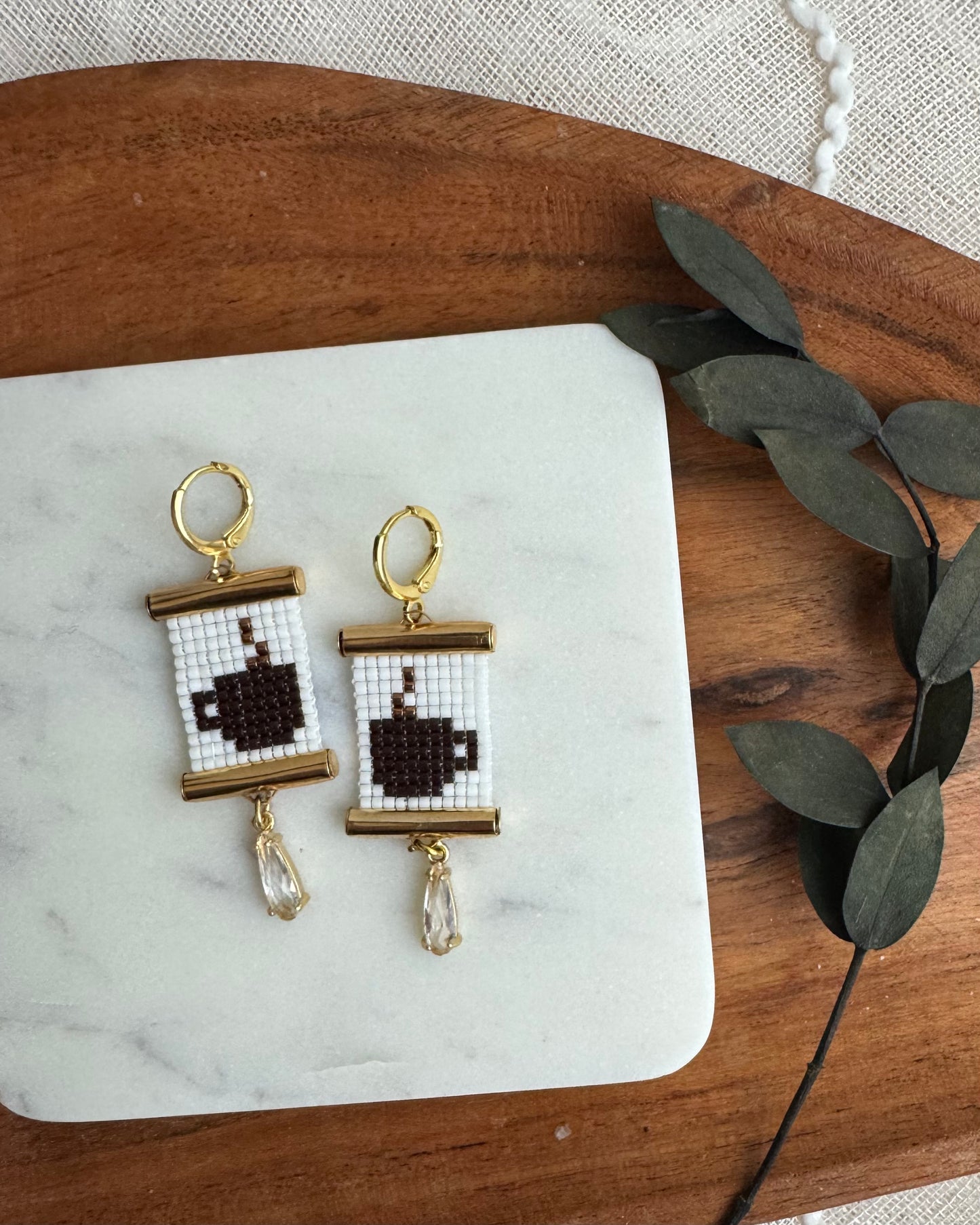 Pair of earrings with coffee cup design on a marble surface with wooden and leaf accents.
