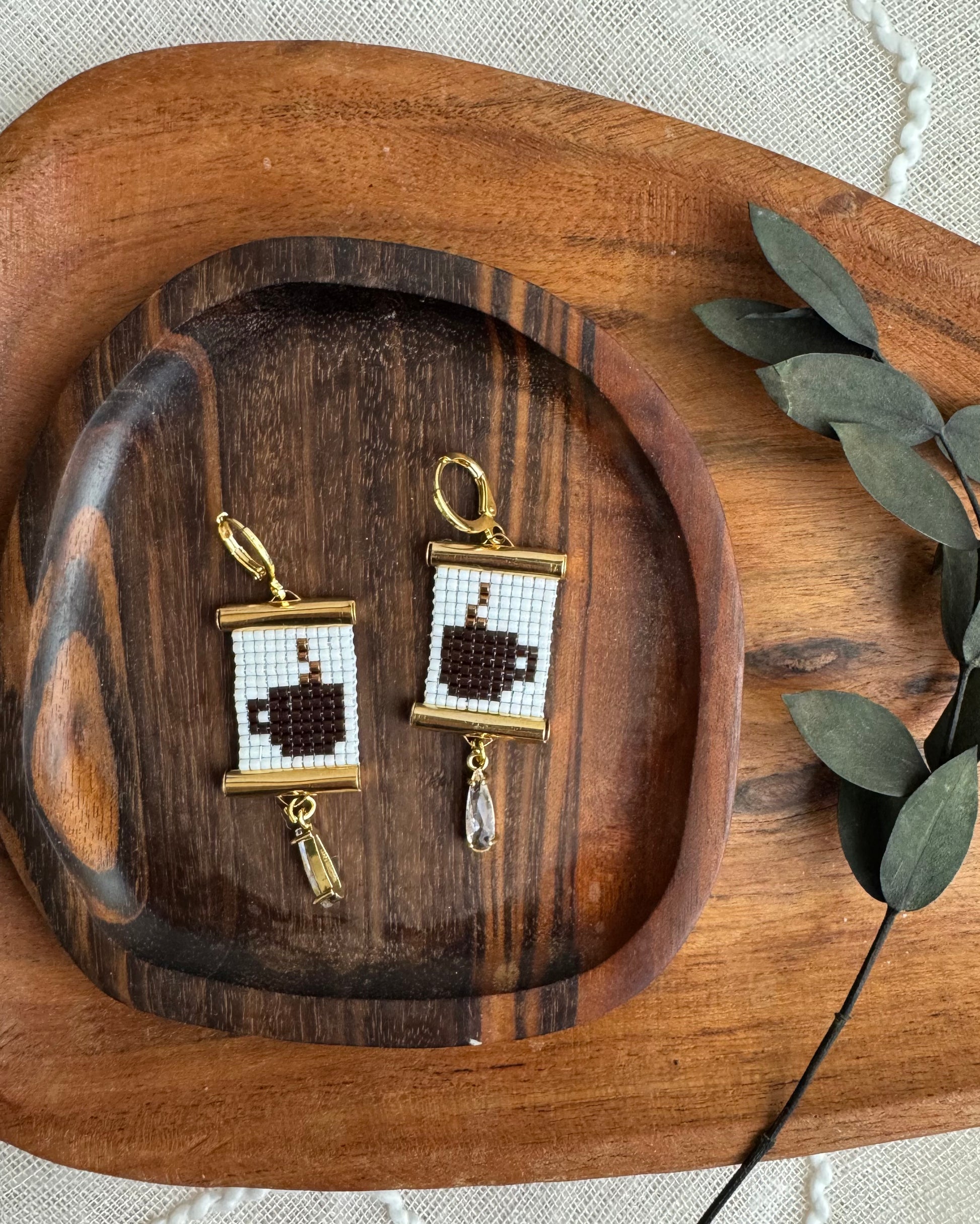 Two gold-framed earrings with coffee designs on a wooden tray.