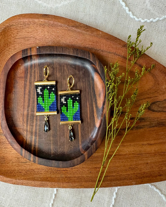 Two beaded earrings depicting saguaro cacti with the moon and stars on a wooden coaster backdrop. 