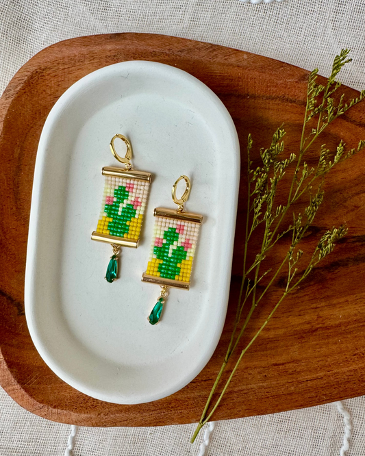 Pair of earrings depicting prickly pear cacti on a white dish with a wooden tray and greenery
