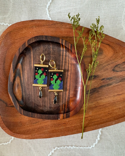 Beaded earrings depicting prickly pear cactii at midnight on a wooden platter with dried flowers.