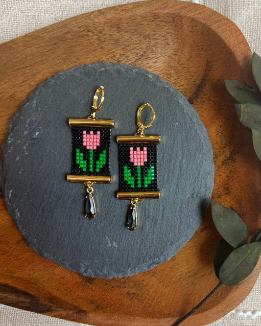 Pair of beaded earrings featuring a tulip design on a slate stone with a wooden base and green leaves.