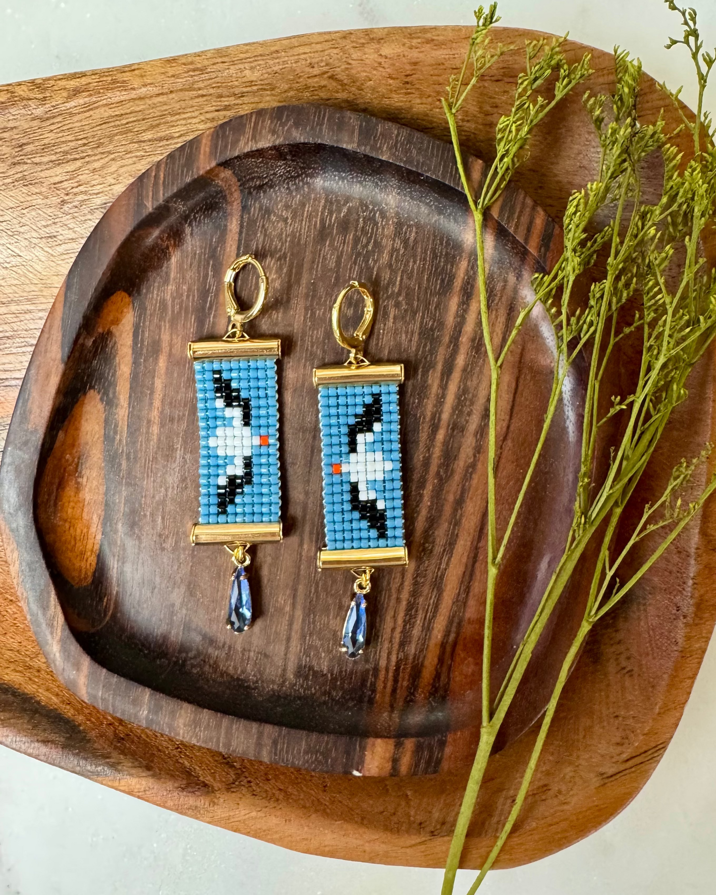 Pair of beaded earrings depicting albatross birds on a wooden tray with a branch on a light background