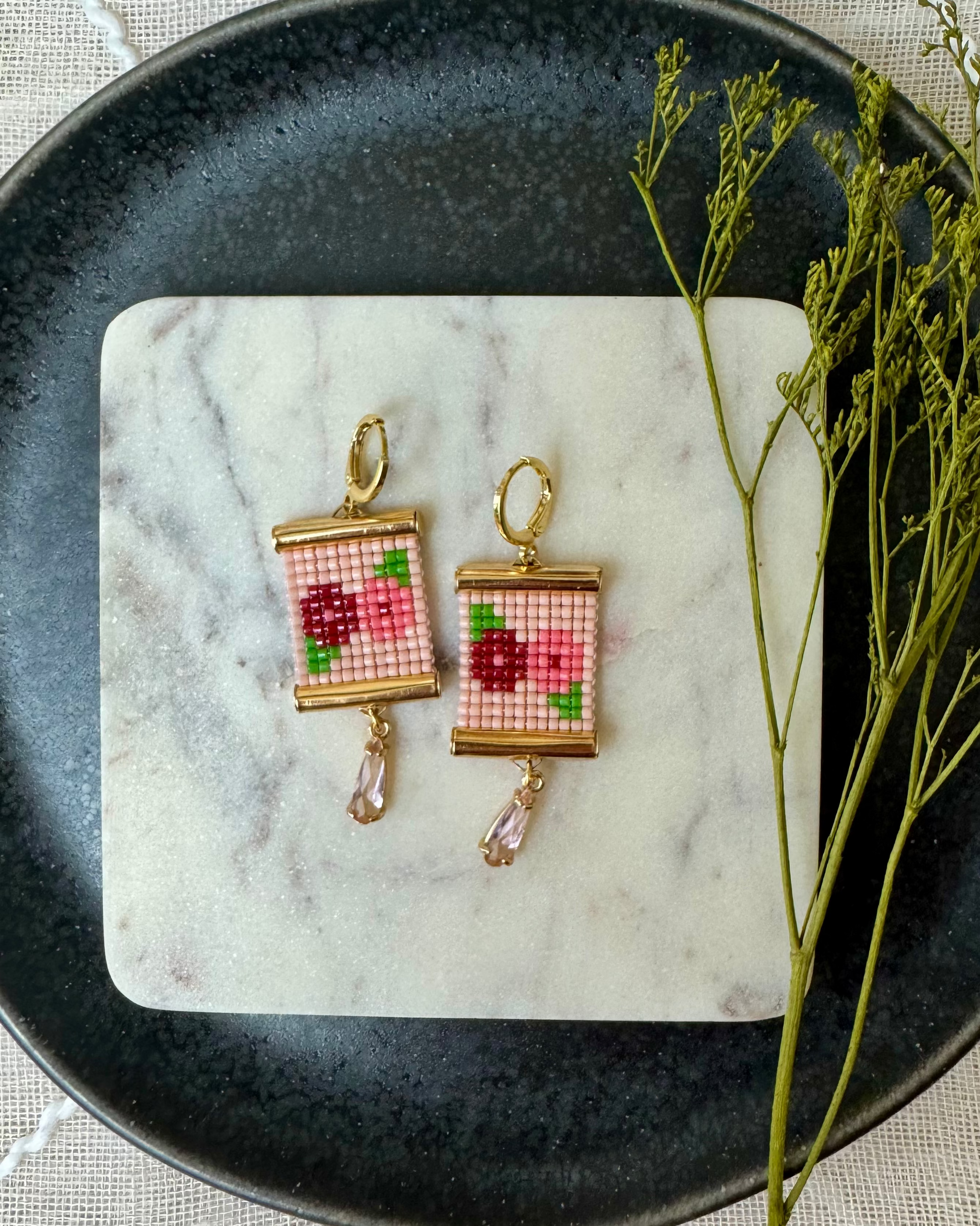 Pair of beaded earrings with flower design on a marble coaster with a black plate and green branch.