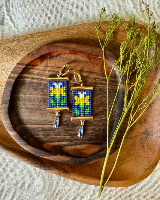 Pair of beaded earrings with daffodil patterns on a wooden tray with dried plants.