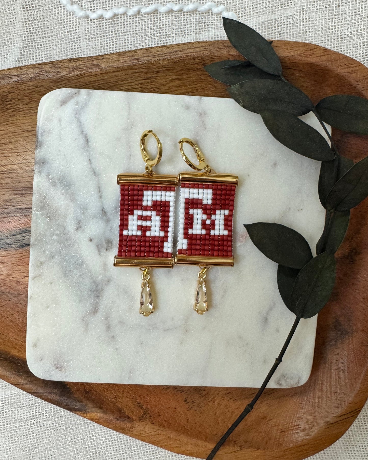 Pair of beaded earrings with red and white design on a marble coaster with leaves, placed on a wooden tray.