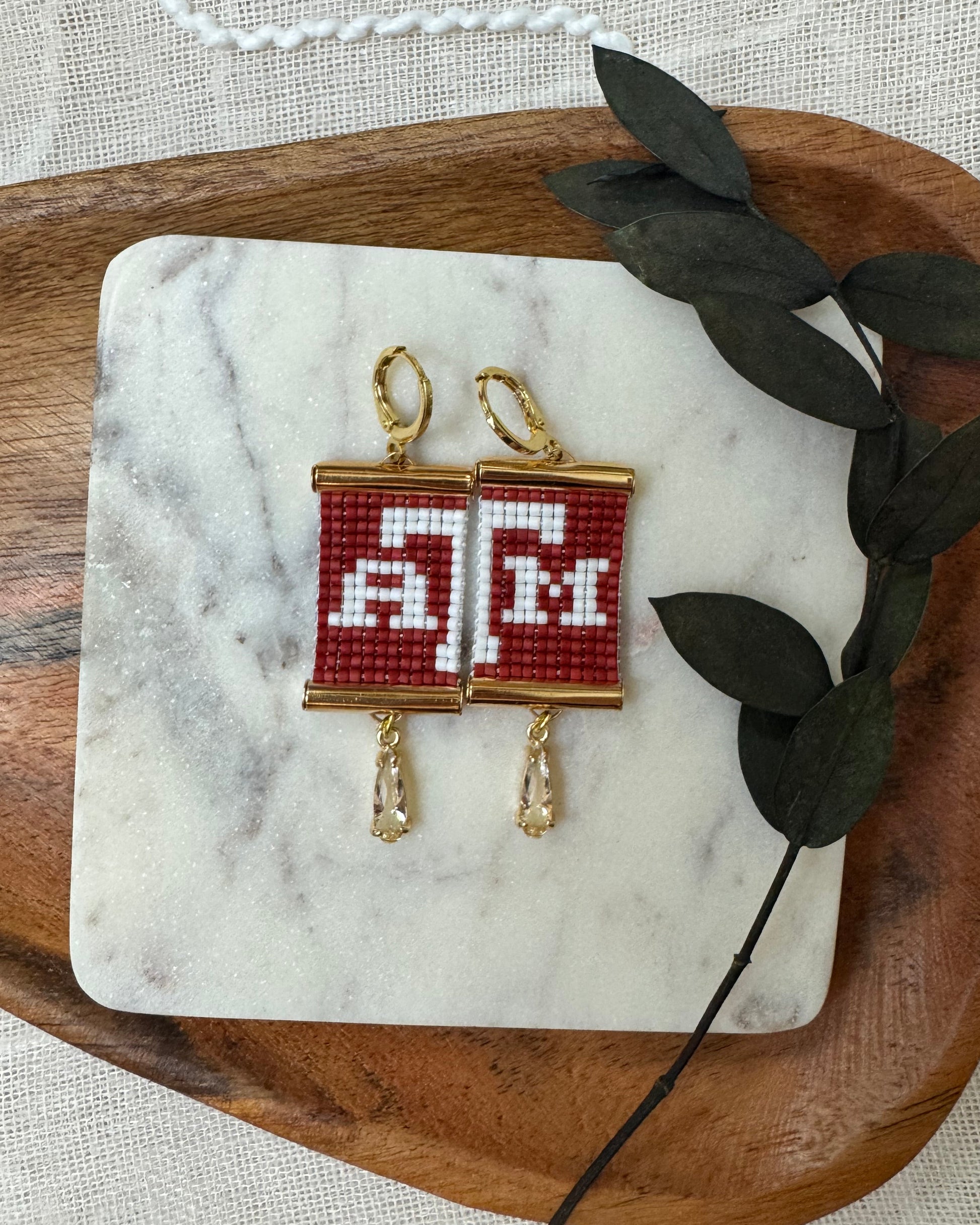 Pair of beaded earrings with red and white design on a marble coaster with leaves, placed on a wooden tray.