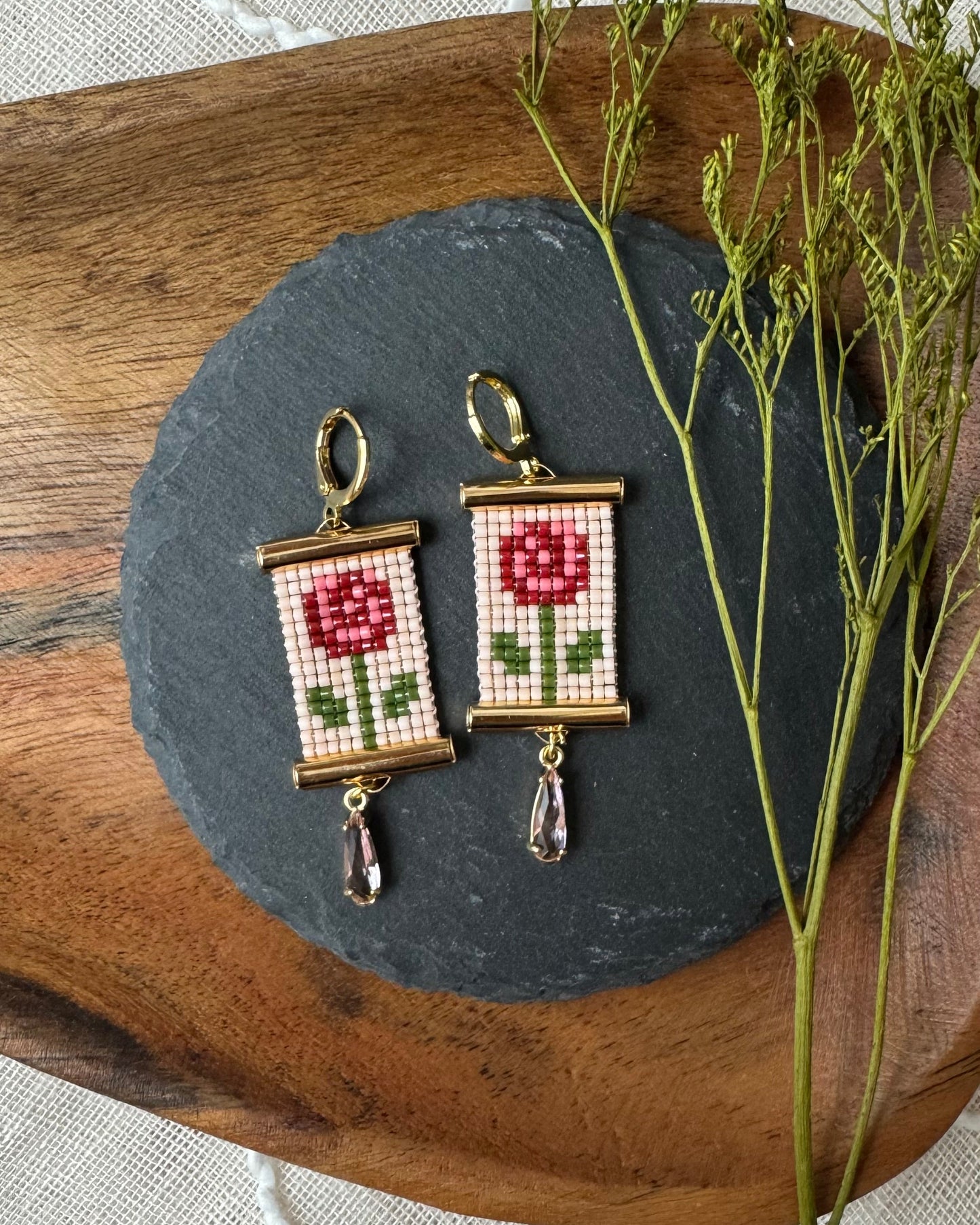 Pair of beaded earrings with rose design on a dark stone surface with a wooden and fabric background.