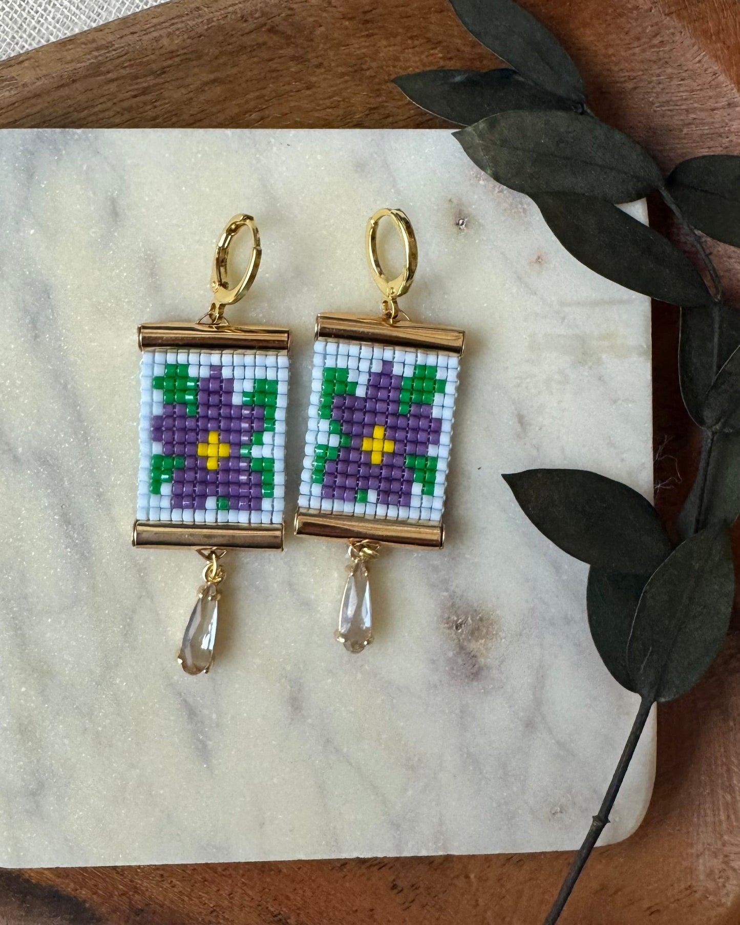 Pair of beaded earrings with floral design on a marble surface with a wooden board and leaf decoration.