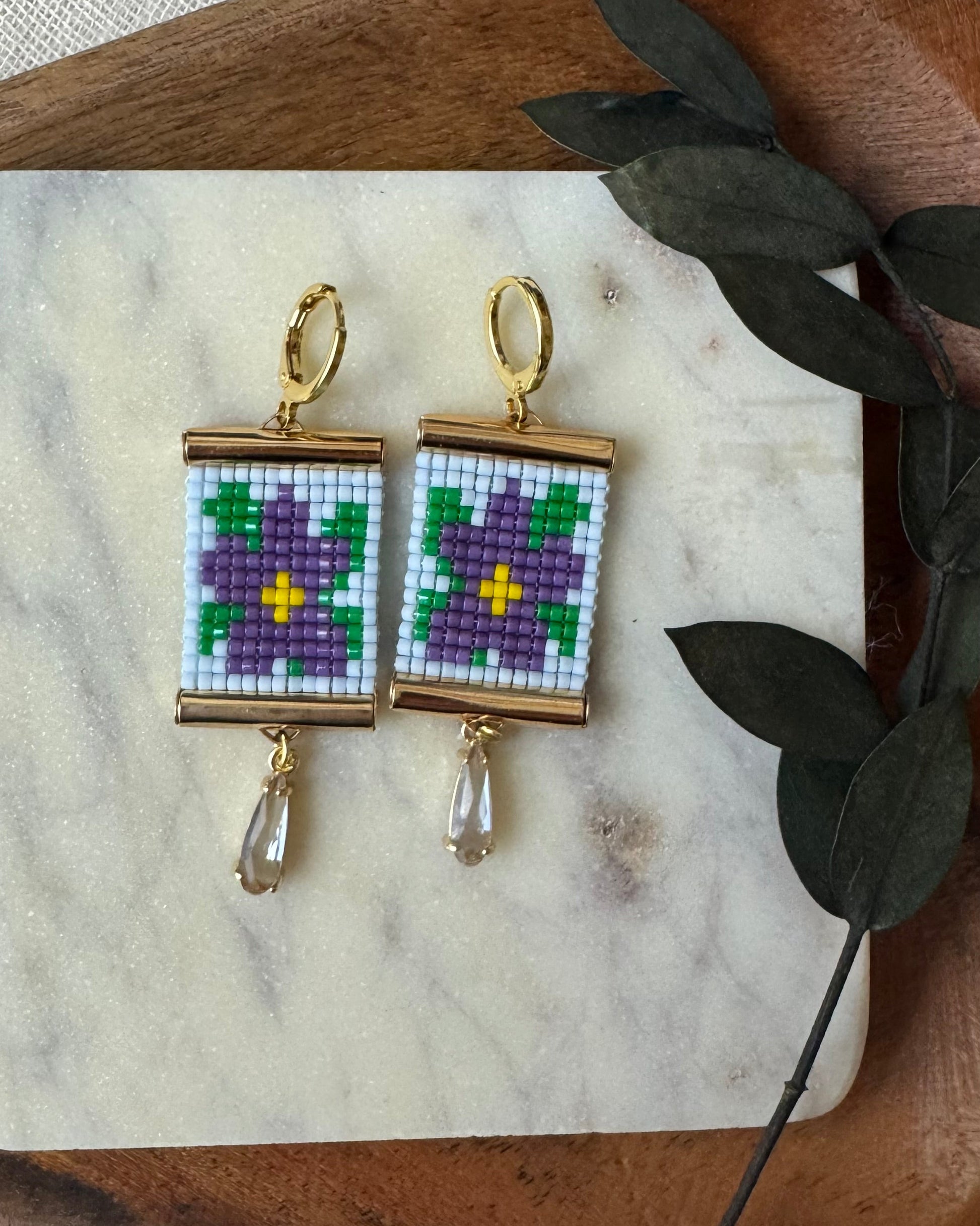Pair of beaded earrings with floral design on a marble surface with a wooden board and leaf decoration.