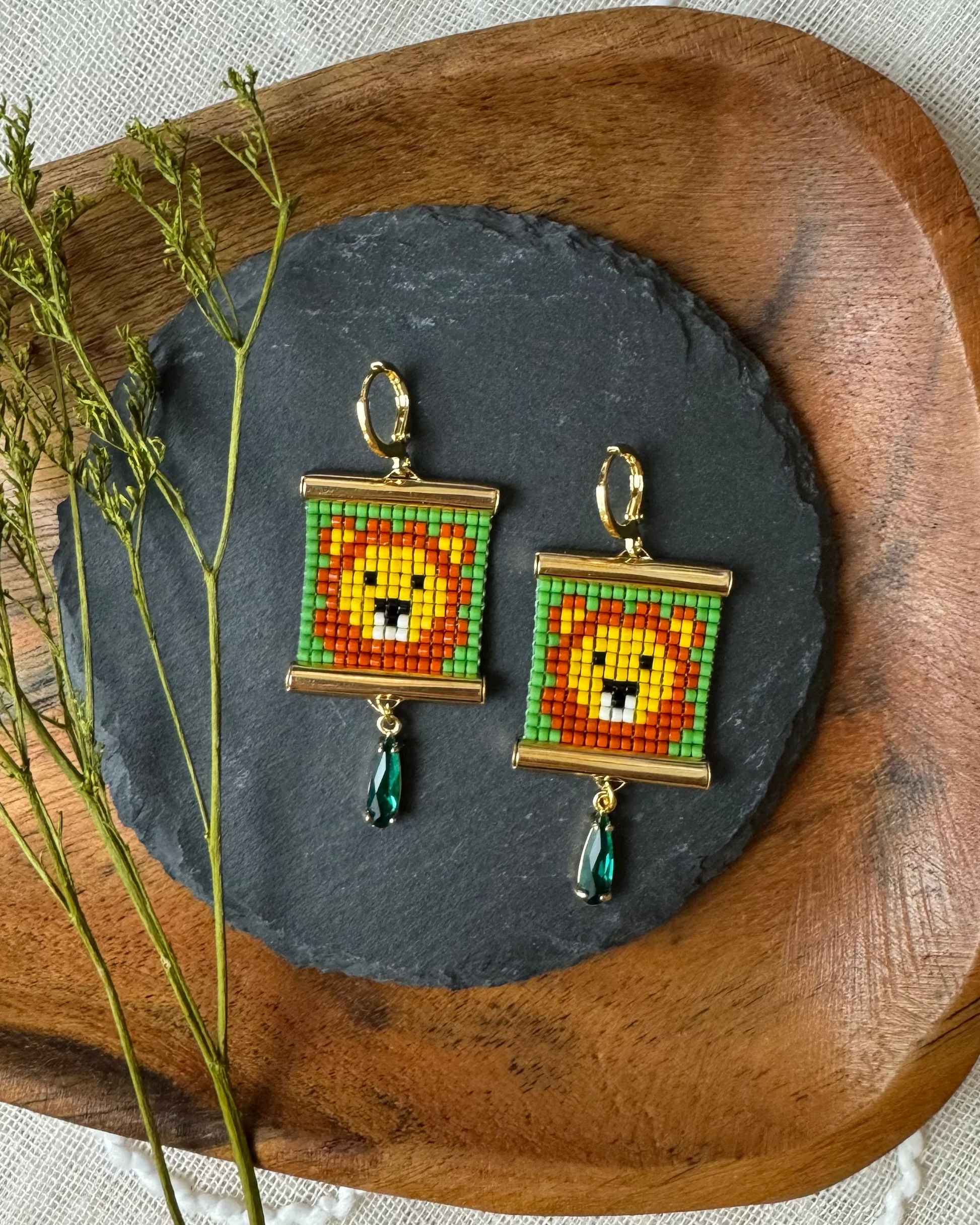 Pair of beaded earrings with lion design on a dark stone surface, surrounded by dried plants.