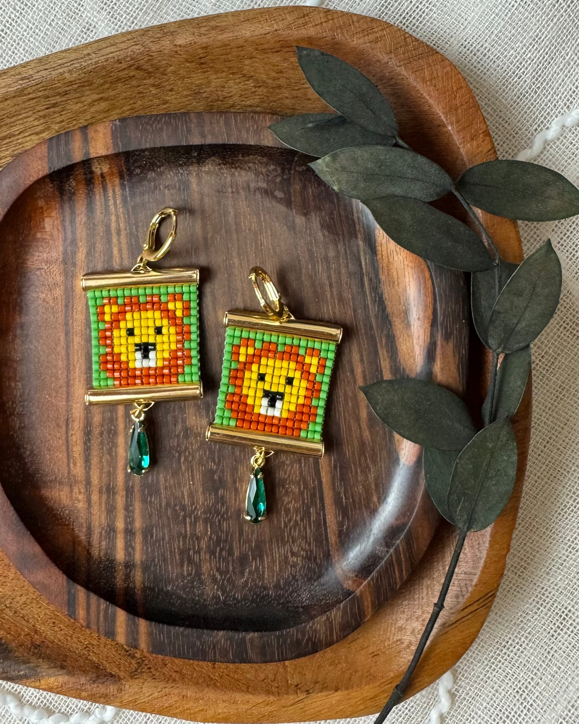 Pair of beaded earrings with lion design on a wooden tray with green leaves.