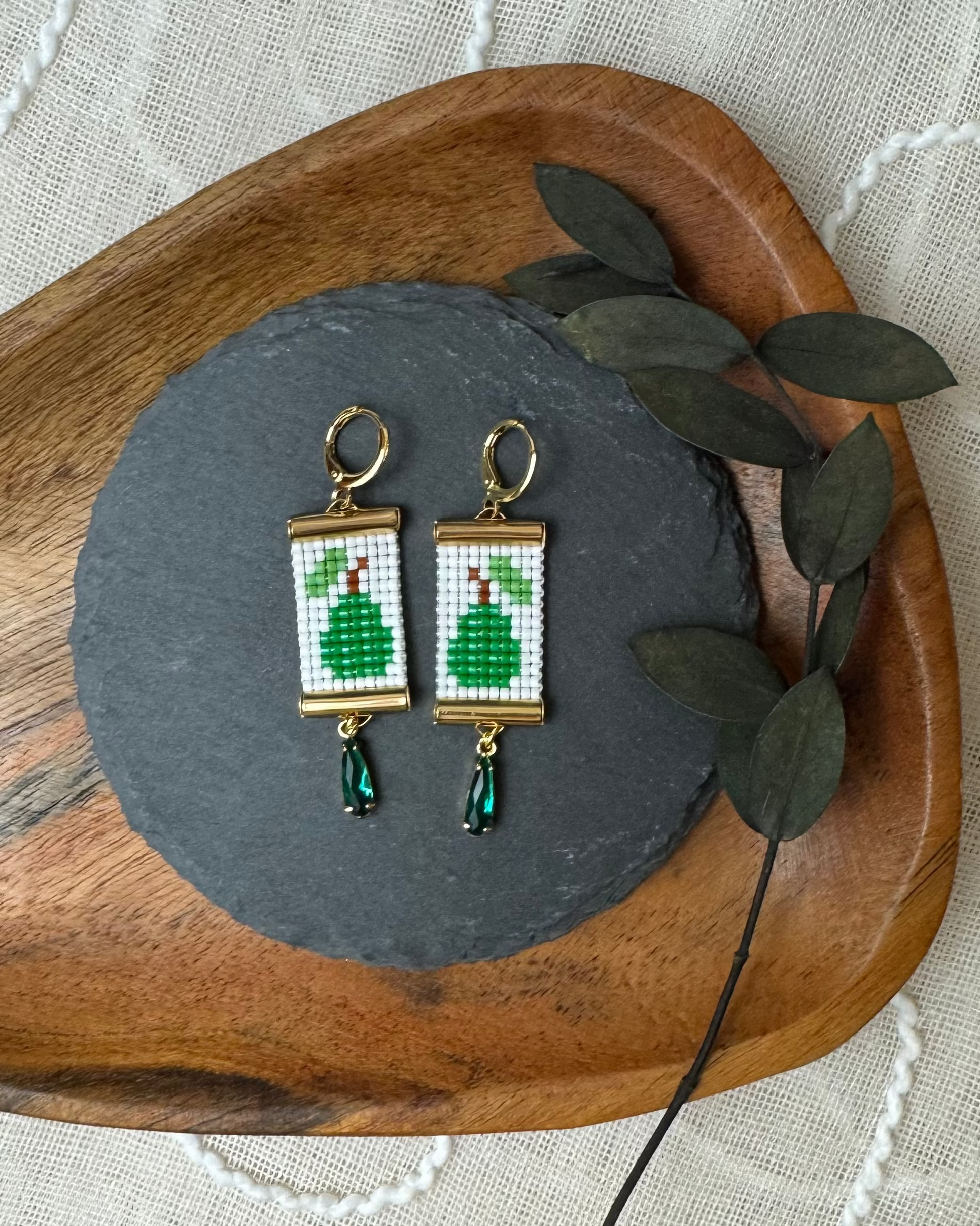 Pair of beaded earrings with pear design on a dark stone surface with a wooden tray and leaves in the background