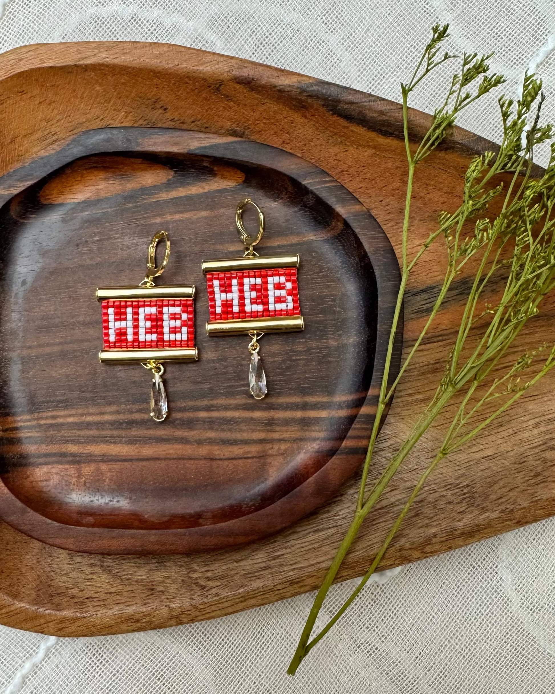 Beaded earrings with 'HEB' text on a wooden tray with a plant.