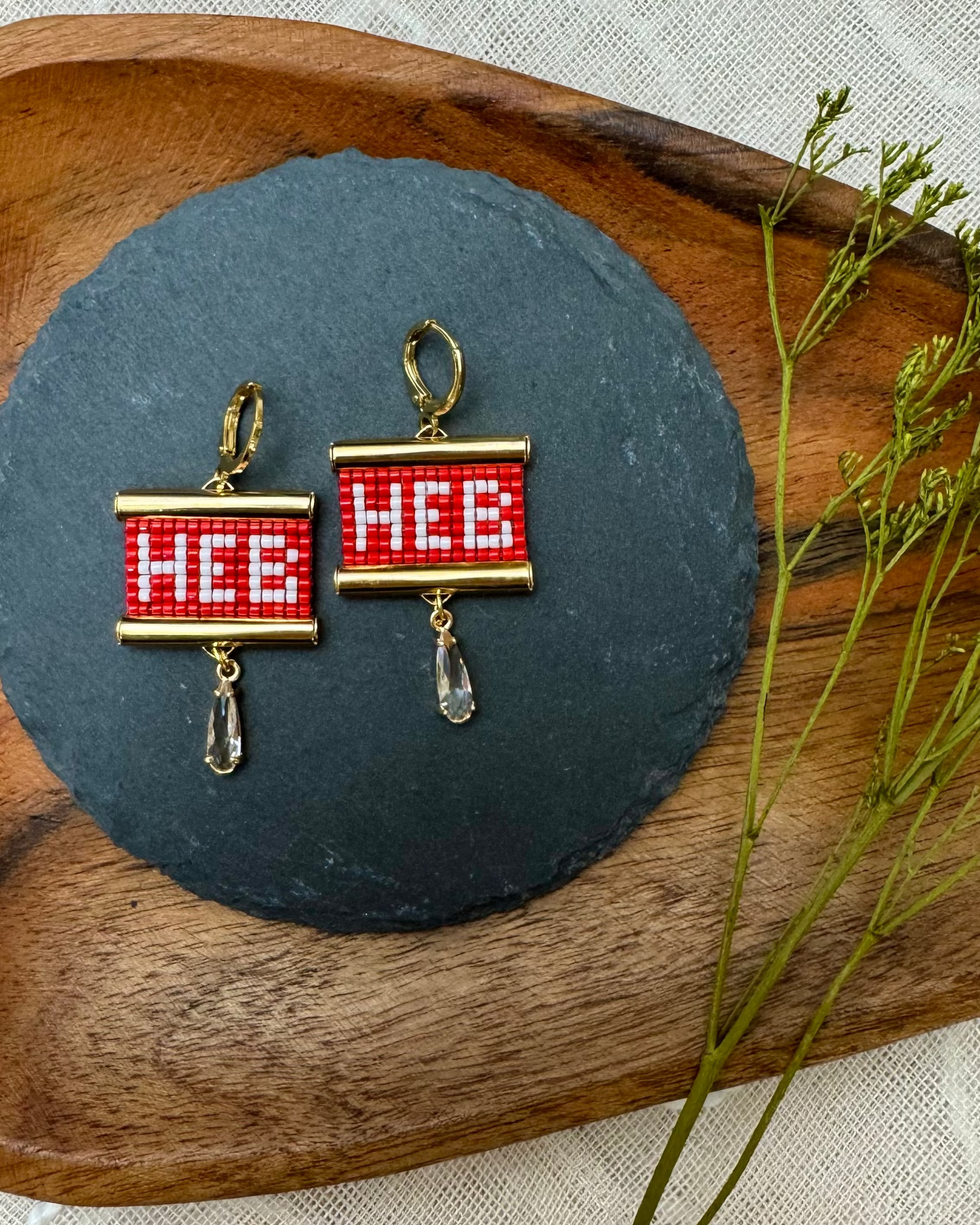 Beaded earrings with 'HEB' design on a dark stone surface with a wooden board and plant in the background.
