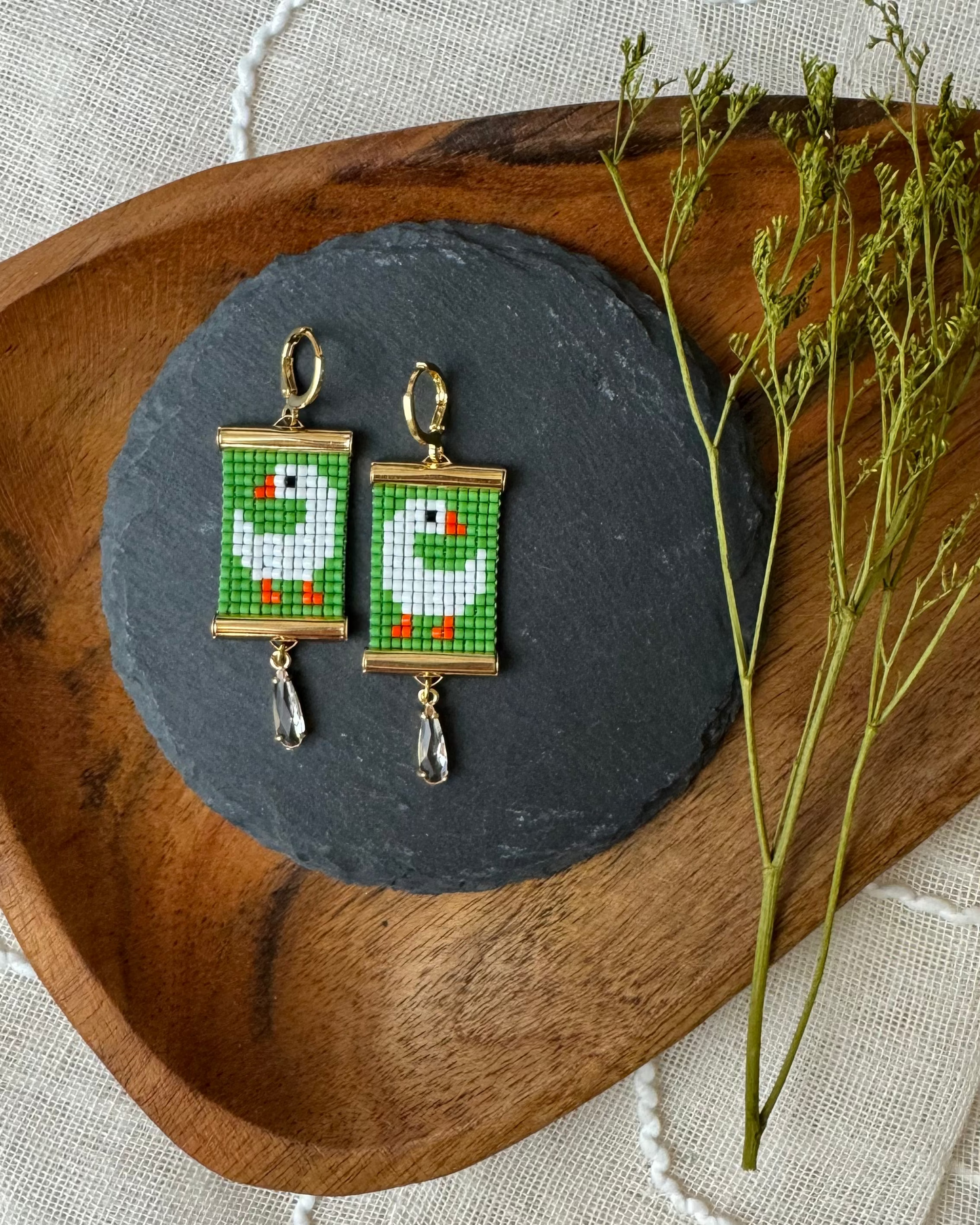 Pair of beaded earrings with goose designs on a dark gray stone surface, placed on a wooden tray.