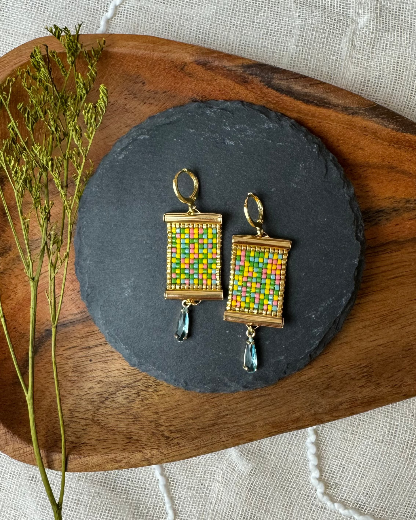 Colorful beaded earrings on a dark slate stone with a wooden board and dried plants.