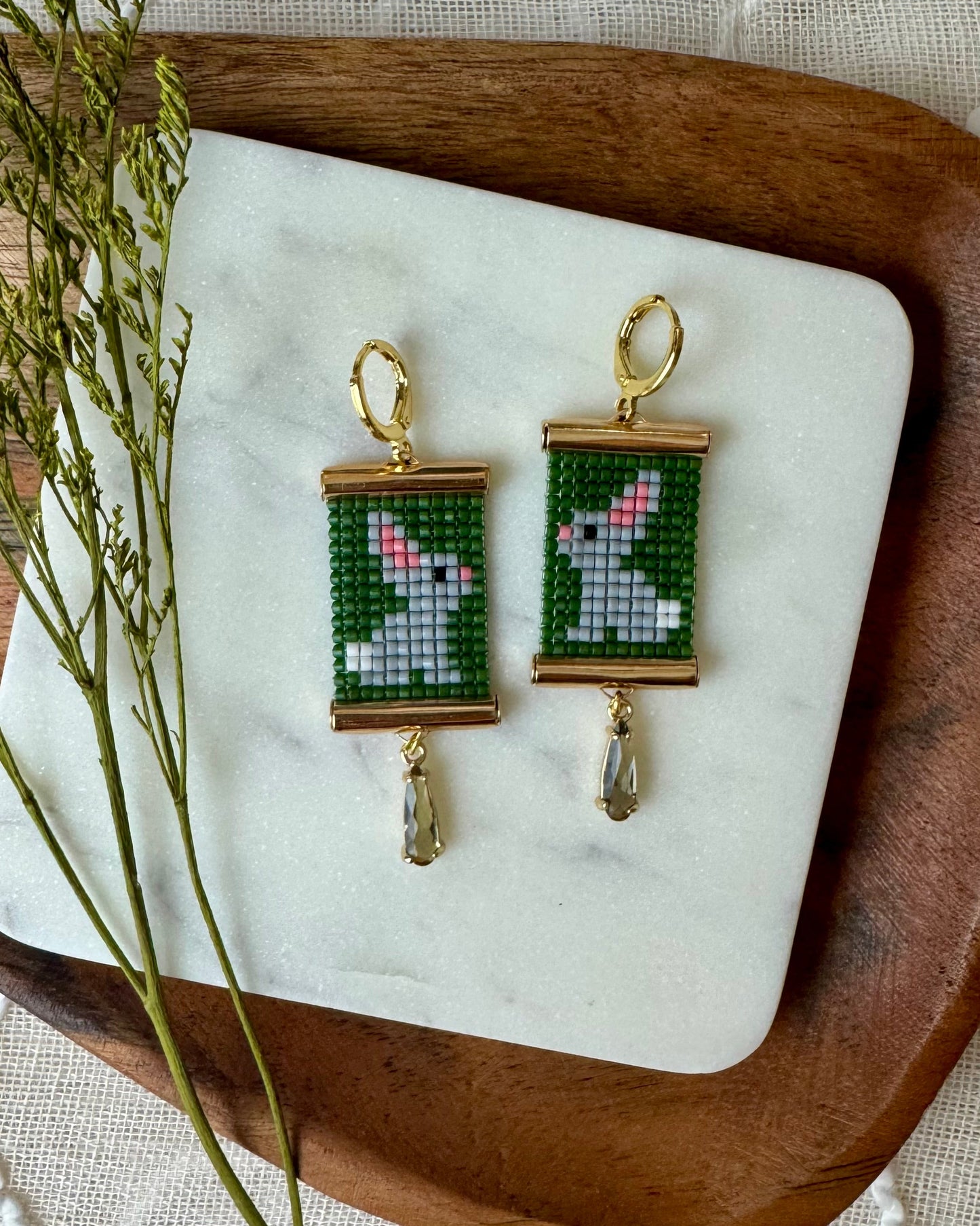Beaded earrings depicting bunnies on a white marble surface with a wooden tray and greenery. 