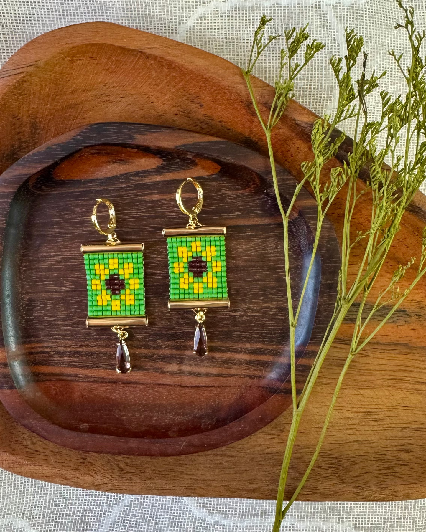 Green and gold beaded earrings on a wooden tray with a plant branch.