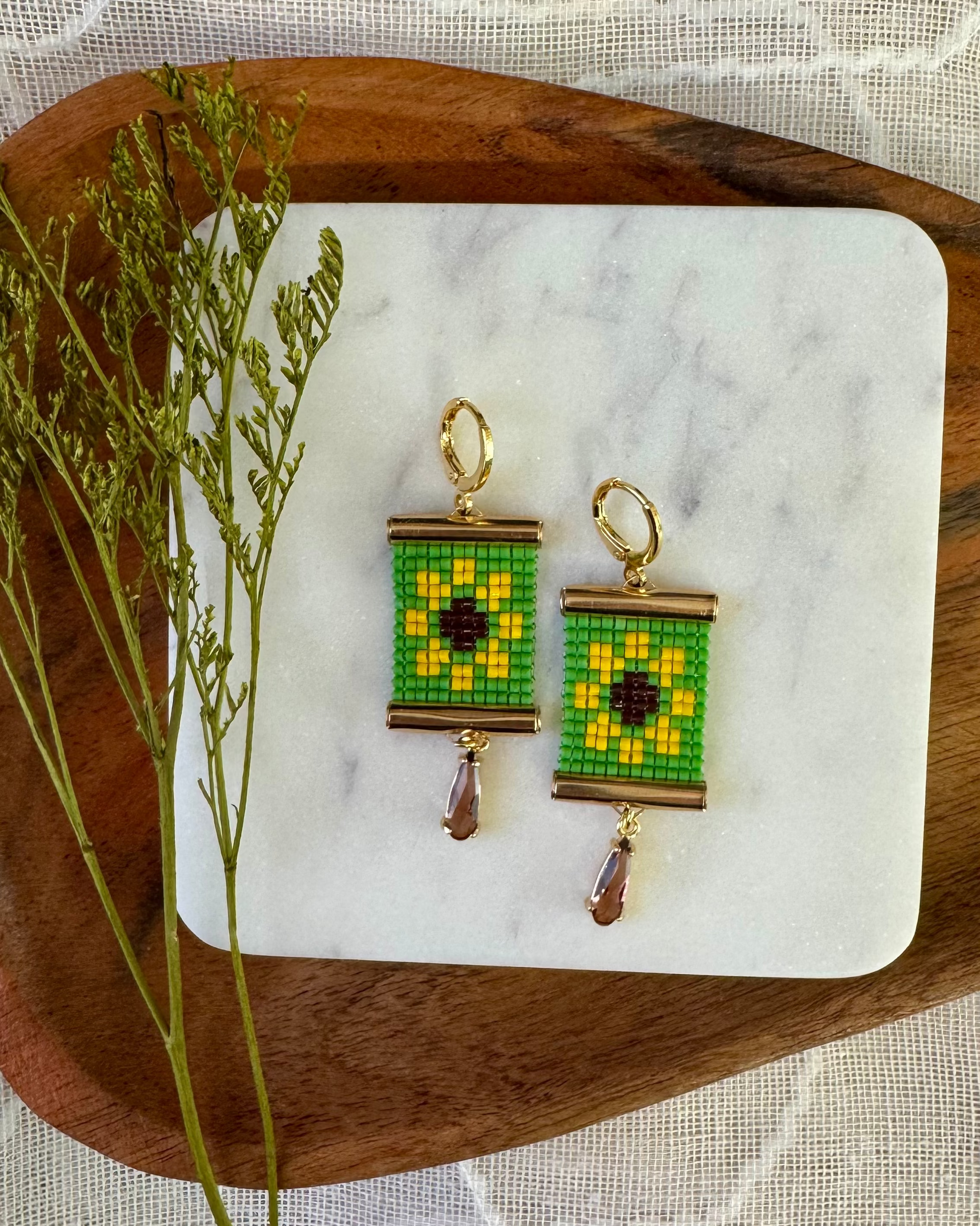 Beaded earrings with green and gold floral design on a marble coaster with wooden tray and plant.
