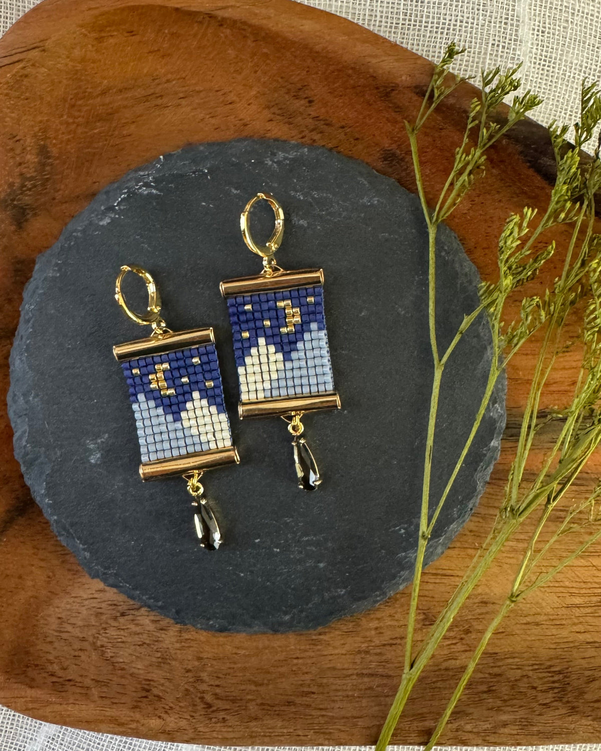 A pair of gold-tone dangle earrings with rectangular blue and white beaded panels featuring crescent moon and star motifs, displayed on a dark slate coaster over a wooden surface with dried green stems.