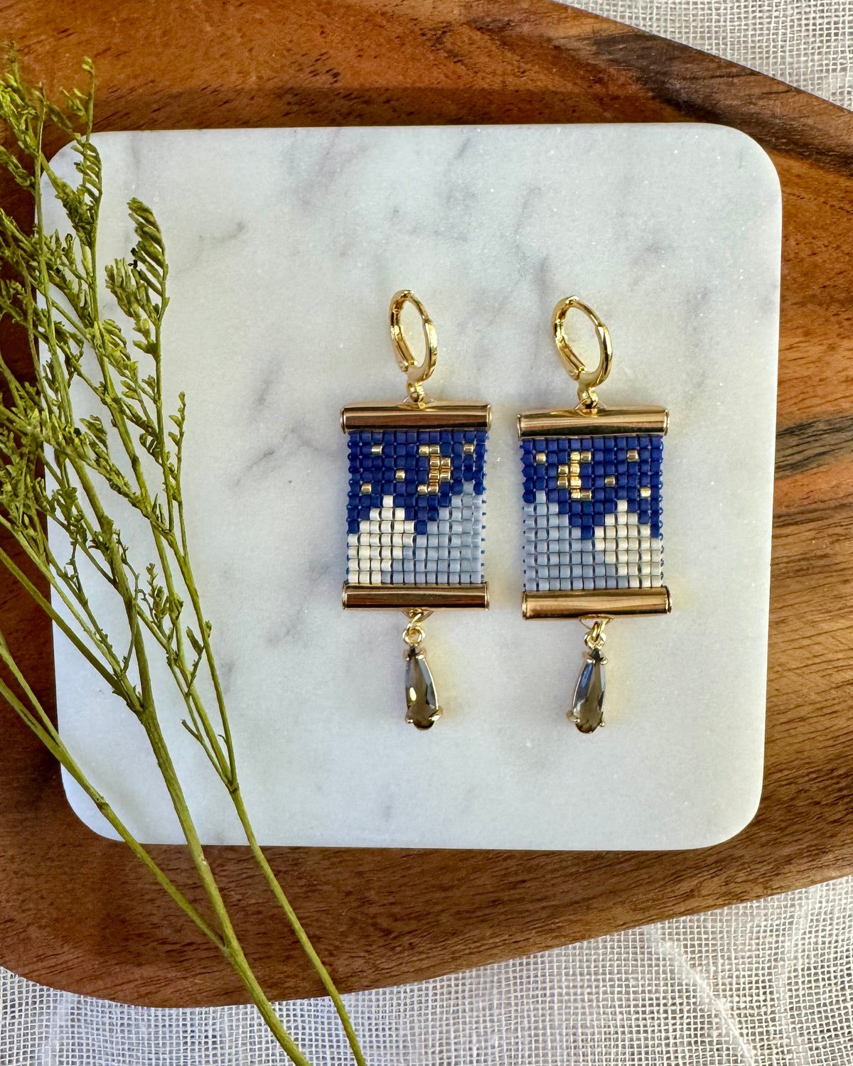 A pair of handmade beaded earrings with blue, white, and gold geometric patterns, displayed on a white marble tray over a wooden surface with green sprigs.