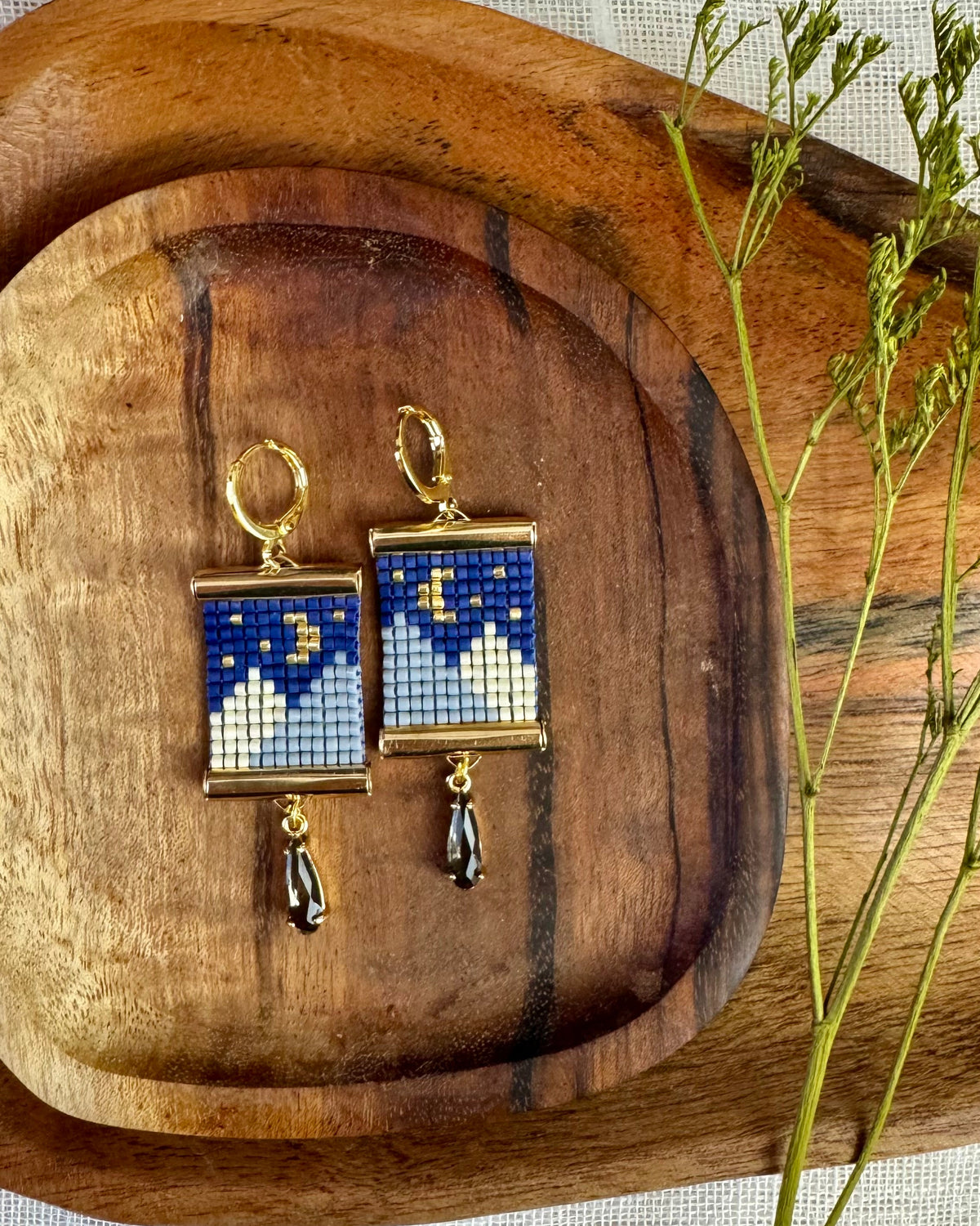 A pair of gold hoop earrings with square blue and white beaded celestial pendants and dangling black teardrop charms, displayed on a wooden tray over a textured white fabric background.